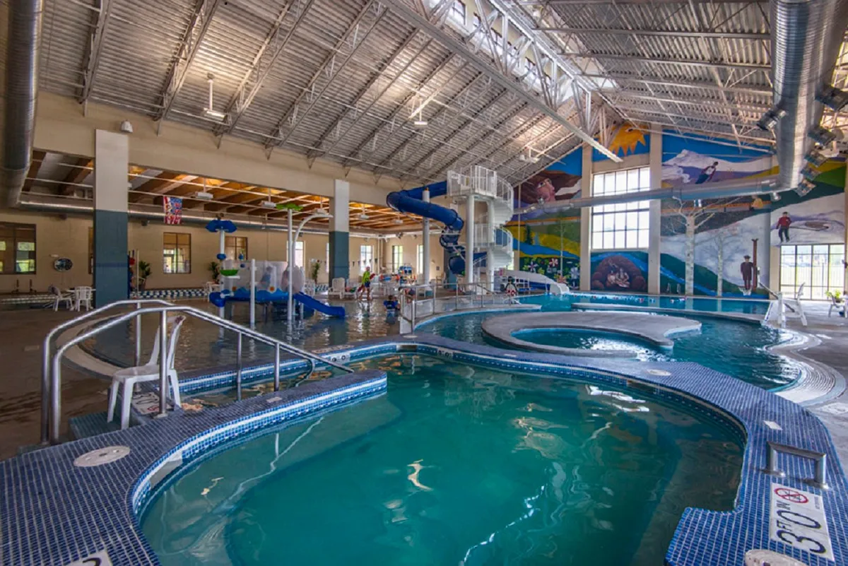 A bustling indoor aquatic center with colorful water slides, a large swimming pool, children playing, and vibrant murals. The joyful, energetic mood suggests a family-friendly environment perfect for year-round fun—indoors and protected from the weather.
