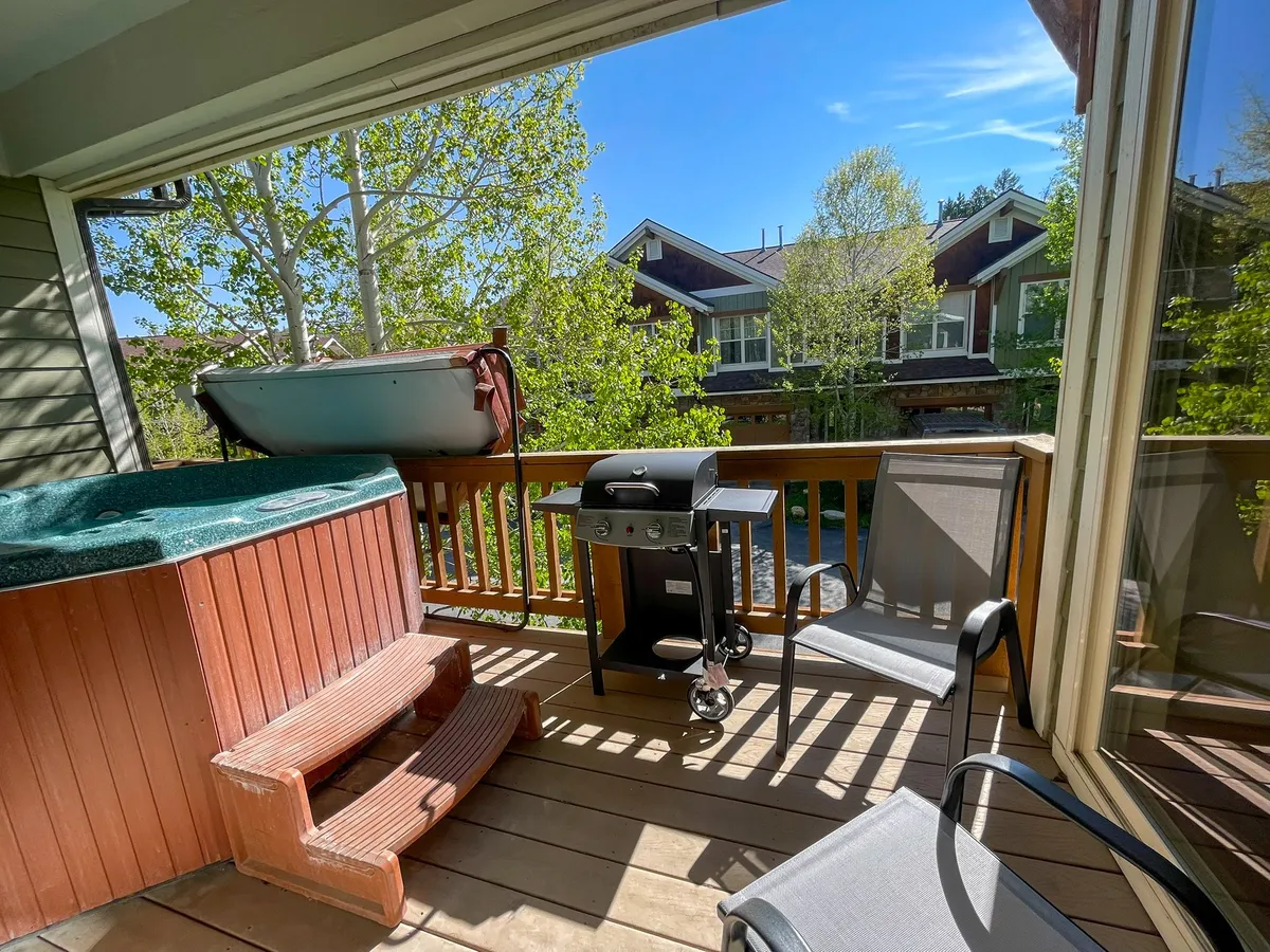 This image captures an outdoor balcony area featuring a bubbling hot tub, an outdoor grill, and comfortable seating. The setting is framed by a scenic alpine backdrop, with the ambiance of early evening suggesting the potential for both relaxation and social gatherings.