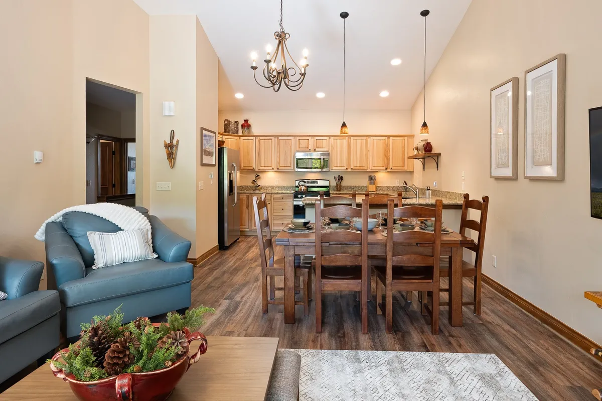 An open-plan interior view displaying the dining table, a cozy sofa, the kitchen area in the background, and an elegant chandelier overhead. The space is set for dining and social interaction, with comfortable lighting and a family-friendly ambiance.