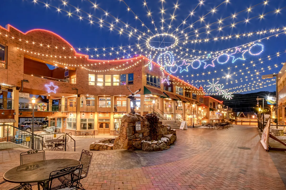 A lively community outdoor area illuminated by string lights with decorative star shapes, a stone fountain, and comfortable seating. The scene evokes a joyful, festive atmosphere, perfect for family strolls, shopping, or socializing.