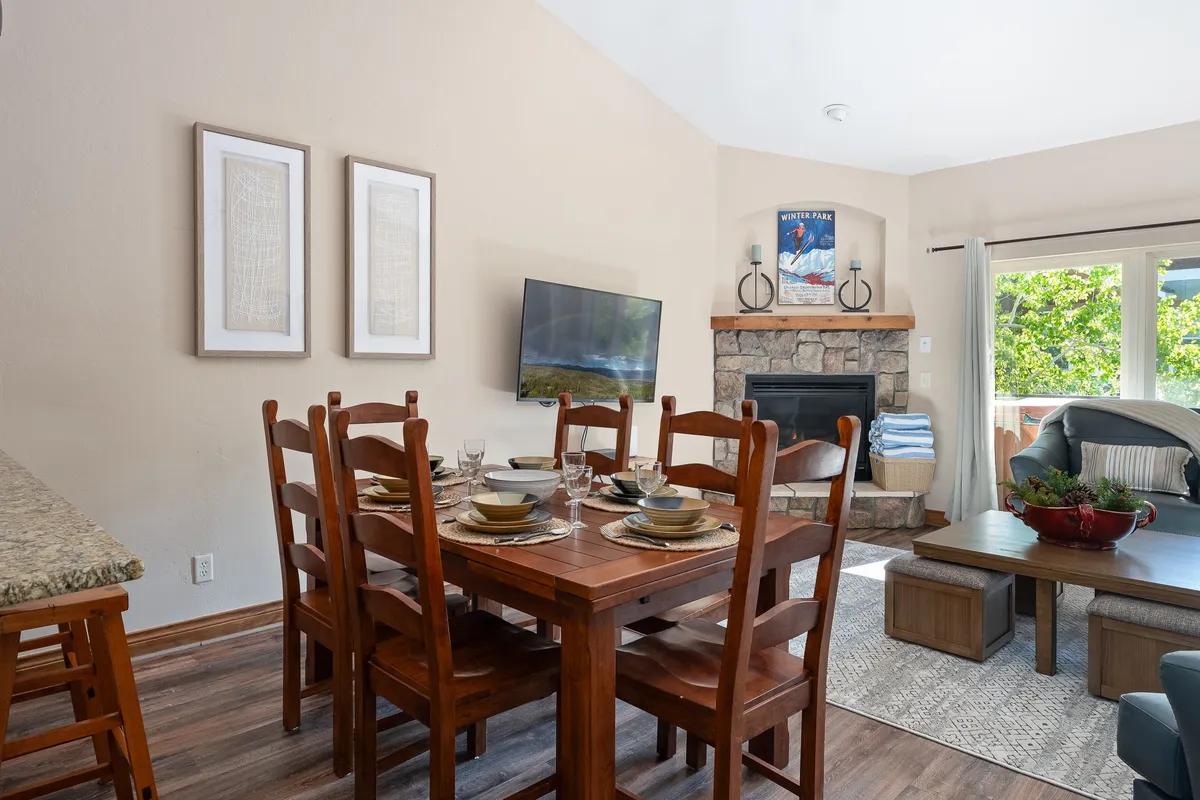 A warmly-lit living and dining area with a dining table set for a meal. A cozy fireplace glows beside a large window, which lets in natural light and offers glimpses of the outdoors. A wall-mounted television is visible, and comfortable seating invites relaxation, setting the stage for family meals, game nights, or unwinding by the fire.