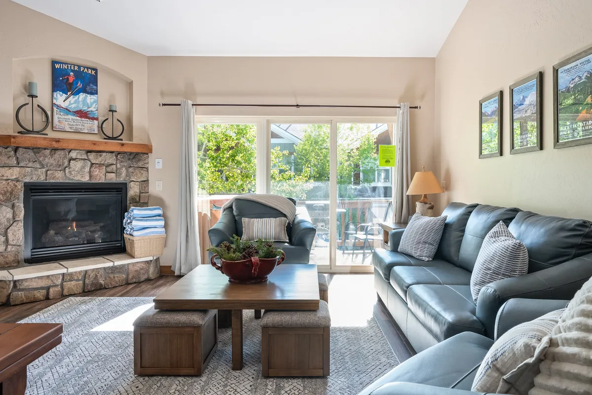 The image showcases a cozy living area with a glowing fireplace, plush seating, a stylish coffee table, and large sliding glass doors leading to a patio with breathtaking sunset views. The space exudes warmth and comfort, inviting guests to relax, socialize, and enjoy the mountain scenery from indoors or step outside for fresh alpine air.