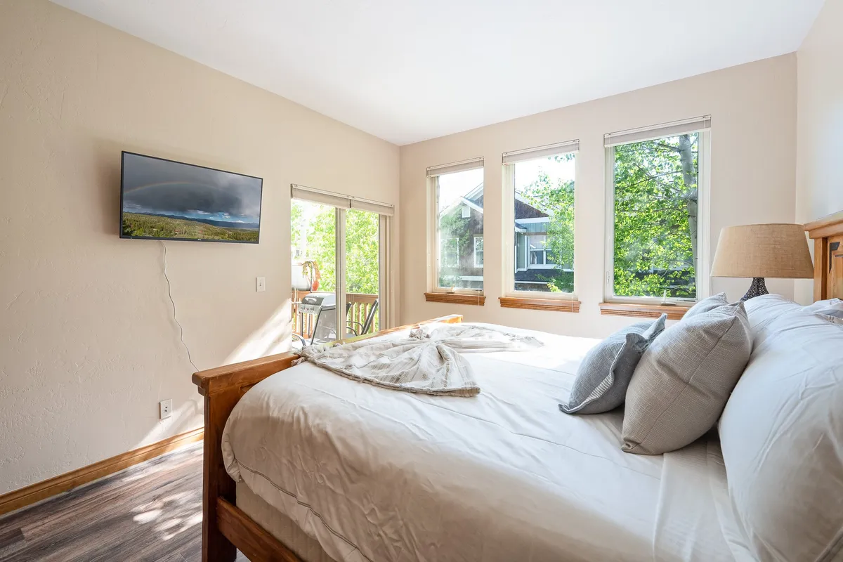A tranquil bedroom featuring a large bed with soft linens, windows that let in ample natural light, a television, and a bedside lamp. The view outside the windows hints at the scenic surroundings.