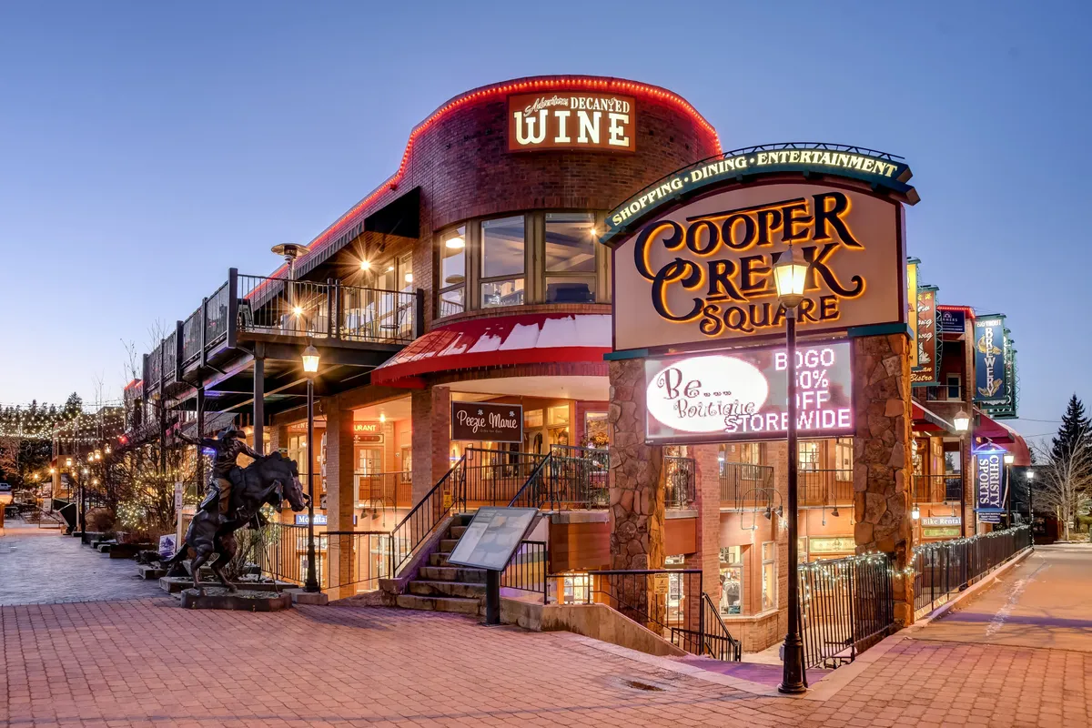 This exterior scene captures the lively Cooper Creek Square, featuring a prominent sign, wine shop signage, outdoor seating, and a staircase leading up to shopping and dining options. String lights and vibrant activity hint at the area's bustling atmosphere.