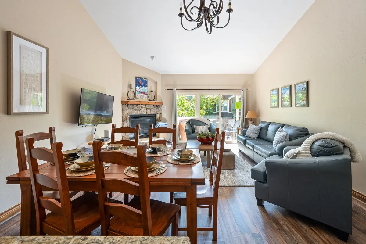A spacious family living area featuring a comfortable sofa, a dining table set for dinner, a glowing fireplace, and a television. Large windows bring in abundant natural light and reveal scenic outdoor views, while the layout encourages both relaxation and group gatherings.