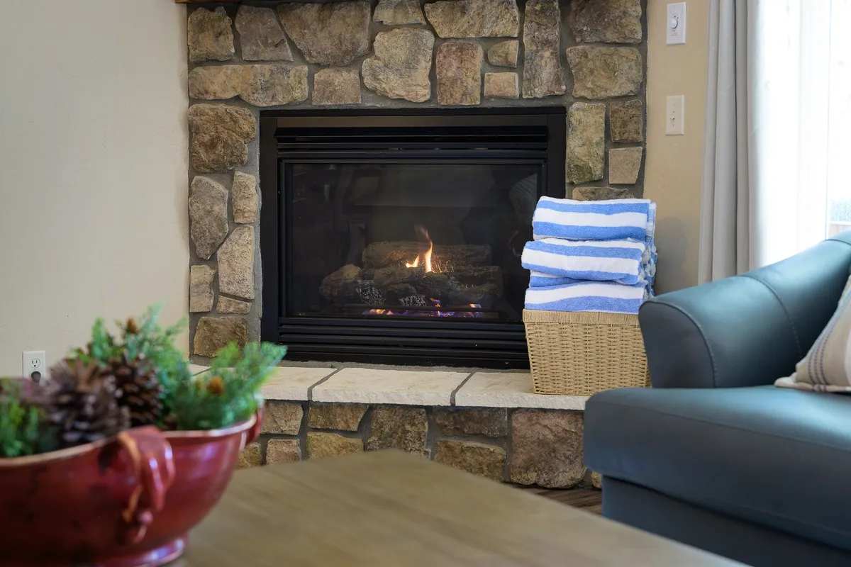 A cozy living room with a stone fireplace, a plush sofa, and an arrangement of greenery. A basket of towels rests by the hearth, and the space is warmly lit, creating a welcoming atmosphere perfect for relaxing after a day outdoors.